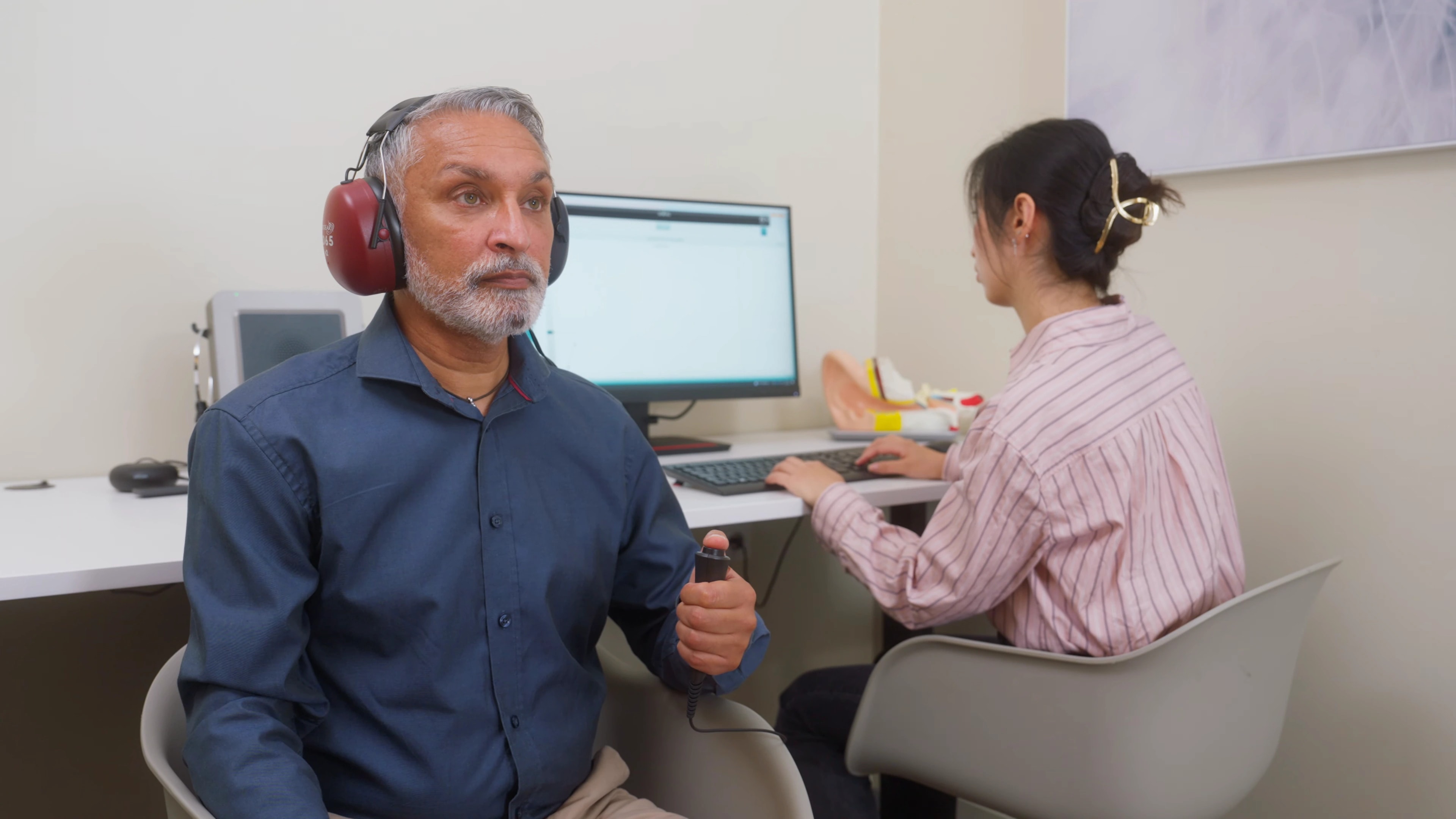 Una persona con auriculares de audiometría sostiene un pulsador de respuesta mientras un operador realiza una evaluación auditiva con Interacoustics.