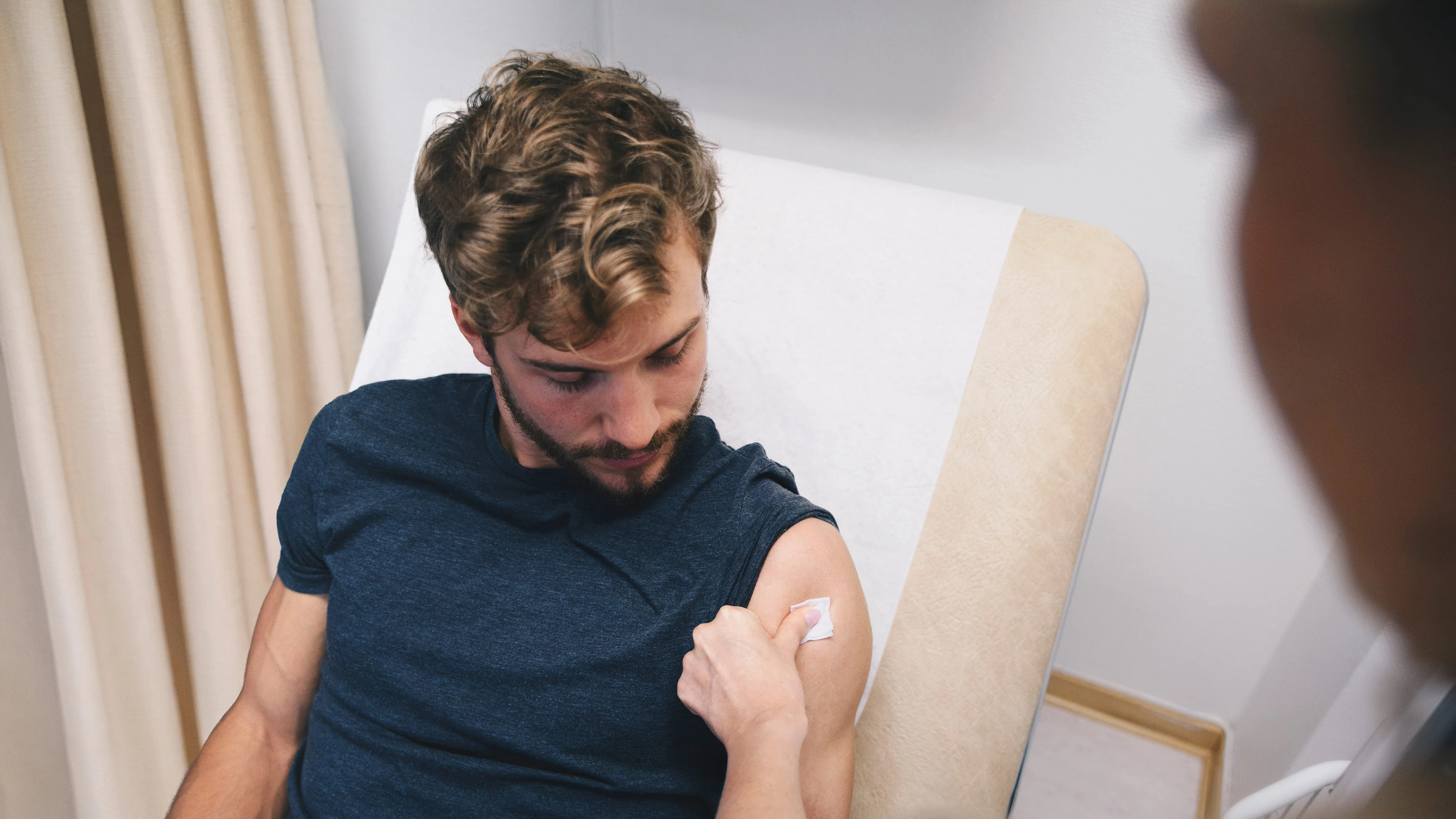 Un paciente presiona un algodón sobre la parte superior del brazo tras una vacunación, con el profesional sanitario en primer plano.